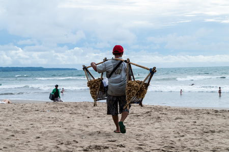 BALI, INDONESIA JANUARY 2017: Peanut seller walks along Kuta beach and tries to sell local peanuts to tourists with heavy load on his shoulders.のeditorial素材