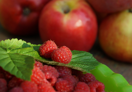 lots of ripe red raspberry berries in a bowl on the table against a background of ripe apples. branch green leaves raspberry on it two berries. summer day in the garden.の写真素材