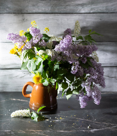 A beautiful still life with lilacs, dandelions, white bird cherry in an old clay teapot on a marble table and a wooden backgroundの写真素材