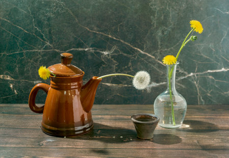 Still life with yellow dandelions in a teapot and a clear glass vase and a white dandelion and an antique lamp on a dark marble background. Conceptual laboratory of life.の写真素材