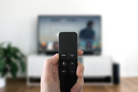 Looking at screen and using remote control. Blurred TV in living room background. Multimedia concept