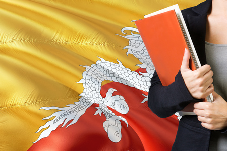 Learning Bhutanese language concept. Young woman standing with the Bhutan flag in the background. Teacher holding books, orange blank book cover.の写真素材