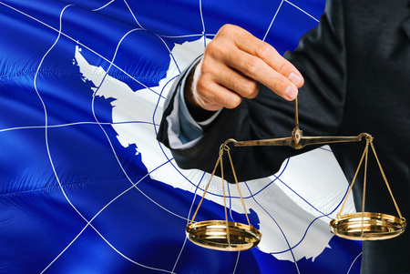Judge is holding golden scales of justice with Antarctica waving flag background. Equality theme and legal concept.の写真素材