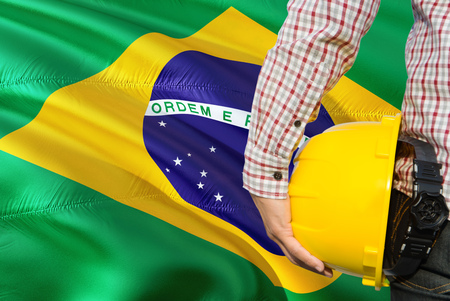 Brazilian Engineer is holding yellow safety helmet with waving Brazil flag background. Construction and building concept.の写真素材
