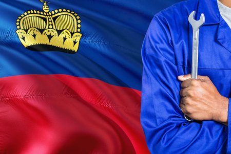 Mechanic in blue uniform is holding wrench against waving Liechtenstein flag background. Crossed arms technician.の写真素材