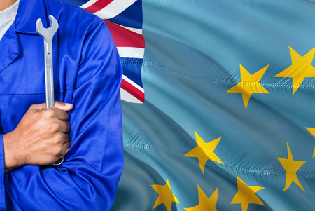 Tuvaluan Mechanic in blue uniform is holding wrench against waving Tuvalu flag background. Crossed...