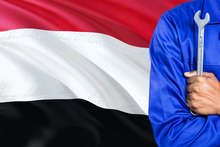 Yemeni Mechanic in blue uniform is holding wrench against waving Yemen flag background. Crossed arms technician.の写真素材