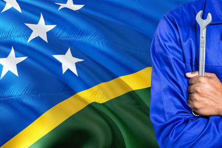 Mechanic in blue uniform is holding wrench against waving Solomon Islands flag background. Crossed arms technician.の写真素材