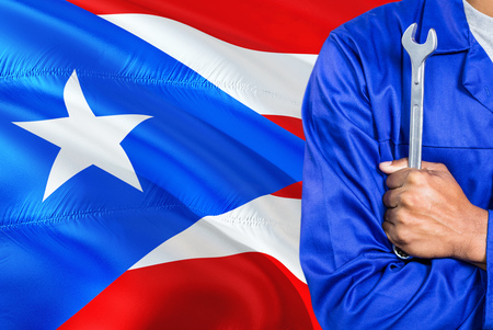 Mechanic in blue uniform is holding wrench against waving Puerto Rico flag background. Crossed arms technician.の写真素材