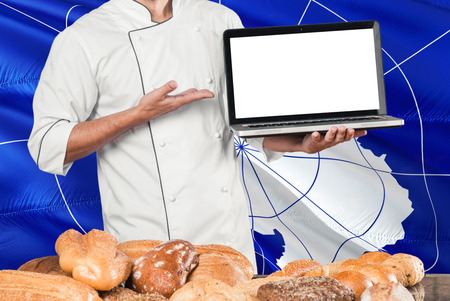 Baker holding laptop on Antarctica flag and breads background. Chef wearing uniform pointing blank...