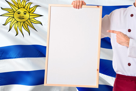 Uruguayan Chef holding blank whiteboard menu on Uruguay flag background. Cook wearing uniform pointing space for text.の写真素材