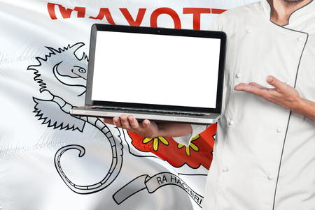 Chef holding laptop with blank screen on Mayotte flag background. Cook wearing uniform and pointing laptop for copy space.の写真素材