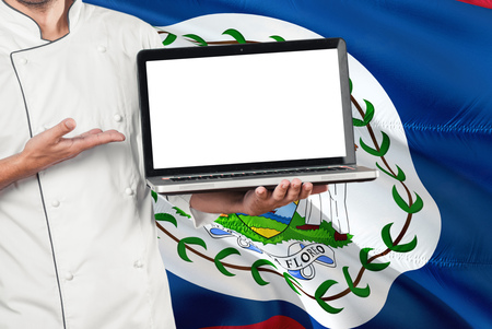 Belizean Chef holding laptop with blank screen on Belize flag background. Cook wearing uniform and pointing laptop for copy space.の写真素材