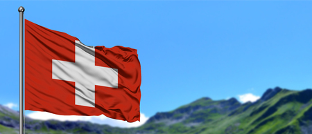Switzerland flag waving in the blue sky with green fields at mountain peak background. Nature theme.の写真素材