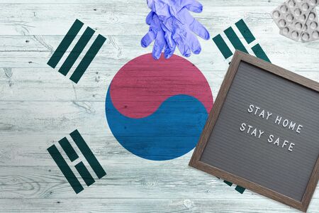 South Korea flag background on wooden table. Stay Home writing board, surgery gloves, pills with...