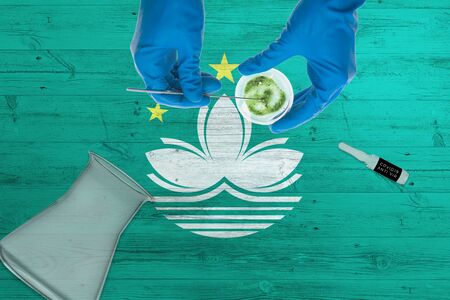 Macao flag on laboratory table. Medical healthcare technologist holding COVID-19 swab collection kit, wearing blue protective gloves, epidemic concept.の写真素材