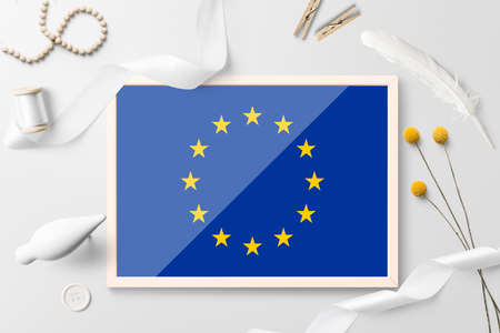 European Union flag in wooden frame on white creative background. White theme, feather, daisy, button, ribbon objects.の写真素材