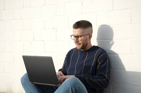 Young caucasian man in casual-wear sitting cross-legged on ground and working on laptop. Freelancer...