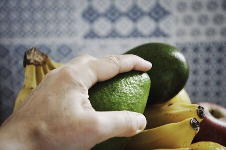 The male hand is taking an avocado from a fruit basket. Fresh green ripe avocado on vintage majolica background. Nutritious and vitaminic food. Healthy food concept. Long life concept.の写真素材
