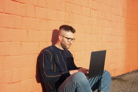 Young caucasian man in casual-wear sitting cross-legged on ground and working on laptop. Freelancer...