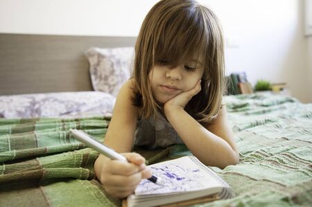 Little cute preschool girl lying on the bed in bedroom at home, she's bored and scribbling with blue...