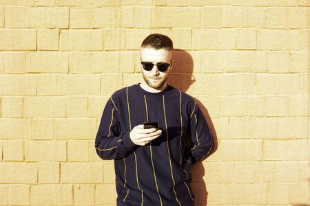 Guy in casual wear with mobile phone - Young caucasian man standing at outdoor red wall and see...