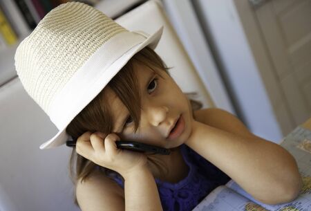 A bored little girl talking with cell phone. Portrait of the cute little girl in casual summer wear and white hat with smartphone. Childhood and tech concept. Boredom, annoyance in children at home.の写真素材
