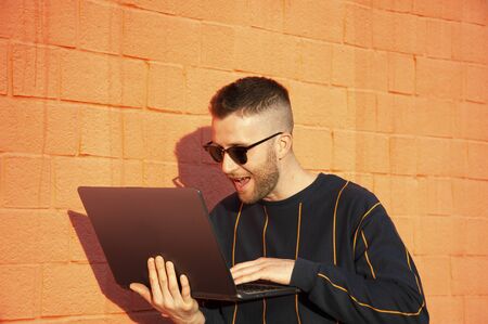 Smiling happy guy looking at laptop monitor, satisfied for result.  Young bearded man enjoying typing on laptop while standing against red wall background outdoors.の写真素材