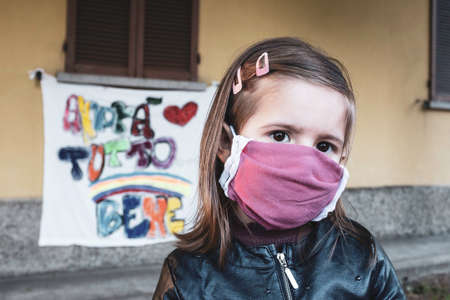 Little girl wearing a protective mask standing in front of the closed school or home during...