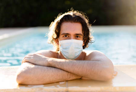 Young man with medical protective mask leaning at the poolside with his arms crossed during coronavirus outbreak - Guy in protective mask inside the swimming pool - Covid-19 impact on travel industryの写真素材