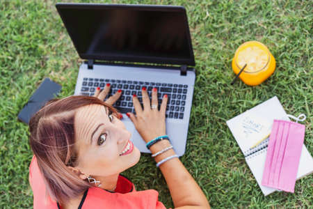 Woman working outdoors with laptop - Smiling female entrepreneur sitting on lawn typing on pc during...