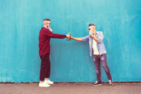 Guys friends toasting beer bottles with opened face masks against blue wall background - New normal lifestyle concept with people having fun together drinking but with social distancing - Copy spaceの写真素材