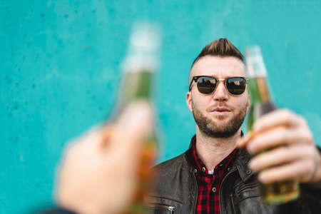 Bearded cool man toasting beer with best friend outdoor against a blue wall background - Hands of hipster man wearing leather jacket toasting with beer bottles - Friendship concept, focus on man faceの写真素材