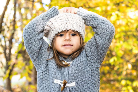 Pensive little girl wearing sweater and wool hat and posing outdoors against autumnal yellow leaves background - Portrait of cute little girl with hands on head during foliage time at park or forestの写真素材