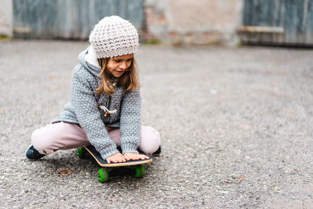 Adorable little girl in winter wool clothes sitting on skateboard outdoors in backyard - Smiling little girl playing with skateboard - Playful childhood and healthy sport conceptの写真素材