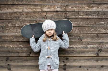 Portrait of caucasian little girl athlete skateboarder in wool winter clothes with the skateboard on his shoulders looking at camera on wooden background - Childhood and active sport conceptの写真素材