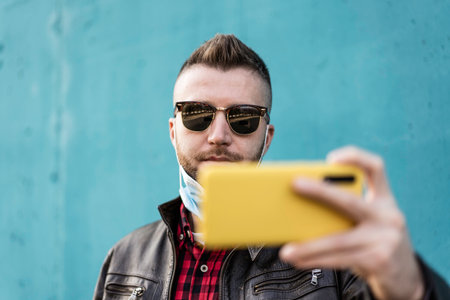Cool man with yellow cellphone wearing open face protective mask during virus outbreak - Hipster man video calling against blue wall background - Tech and new normal concept during pandemic timeの写真素材