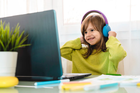 Elementary schooler girl in headphones sitting at desk on laptop - Smiling cute girl studying online...