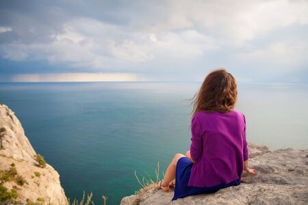 Young beautiful girl sitting on a rock and looking at sea.の写真素材