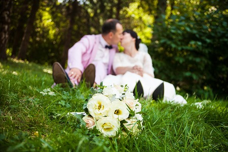 Bride and groom sit on the grass with a beautiful bouquet and a kissの写真素材