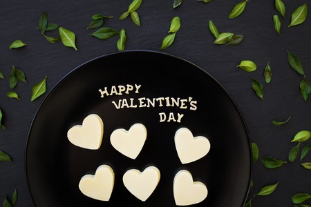 candy white chocolate heart-shaped inscription Happy Valentine on a black plate with green leavesの写真素材