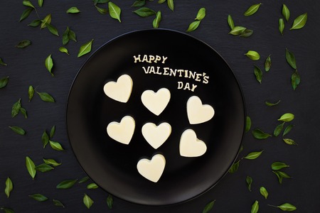 candy white chocolate heart-shaped inscription Happy Valentine on a black plate with green leavesの写真素材