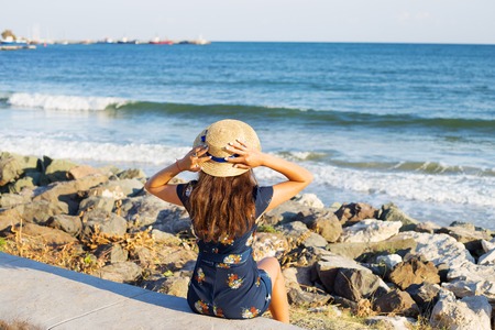 Beautiful girl in a hat sits near the sea on stones.の写真素材