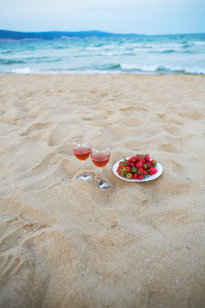 two glasses of wine and a plate of strawberries on a sandy beach, a picnic near the sea.の写真素材