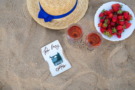 straw hat, two glasses of wine and a plate of strawberries on a sandy beach, close-up.の写真素材