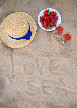 straw hat, wine and a plate of strawberries on a sandy beach-inscription love the sea.の写真素材