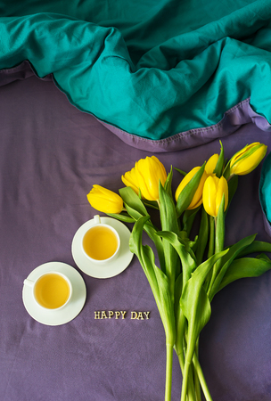 Yellow tulips and two cups of tea in bed, close-up, lettering happy dayの写真素材