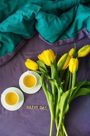 Yellow tulips and two cups of tea in bed, close-up, lettering happy dayの写真素材