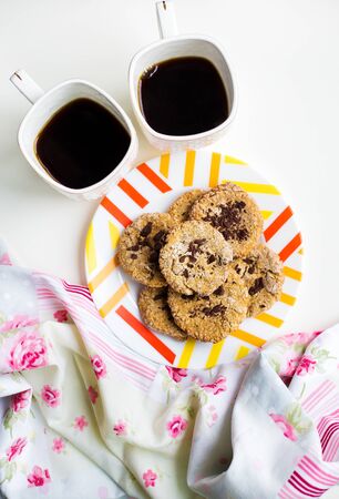 Oatmeal cookies with chocolate on a plate with bright cloth and two cups of coffeeの写真素材