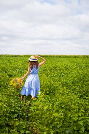 Cute girl in a hat stands in a green field with a bouquet of wild flowers.の写真素材